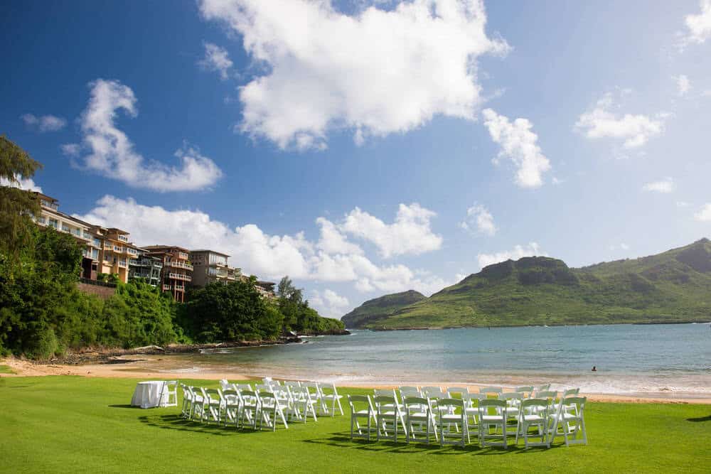 Beach wedding at Royal Sonesta Kauaʻi Resort with ceremony arch on the sand, palm trees, and ocean waves in the background.