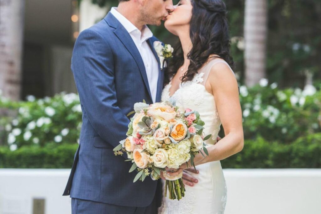 Couple kissing during wedding ceremony designed by Spinning Web Florist, with the bride holding a lush floral bouquet and elegant arrangements surrounding them.