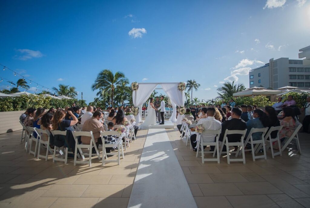 Outdoor Hawaii wedding ceremony at Hyatt Regency Waikiki with the couple beneath a white floral arch, surrounded by guests and palm trees, photographed by Blessed Creatives.