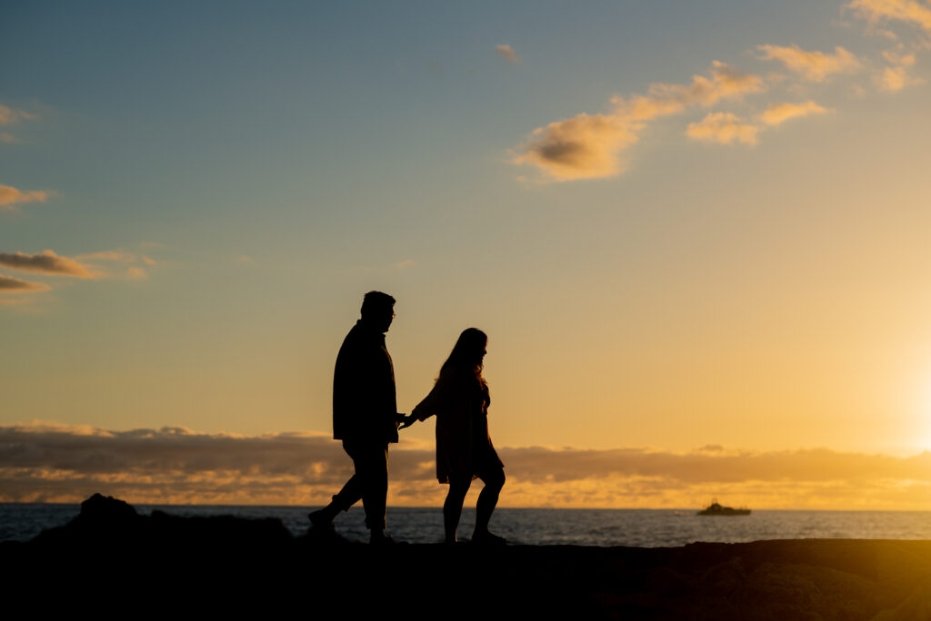 A husband and wife appear as silhouettes while holding hands on a beach during sunset.