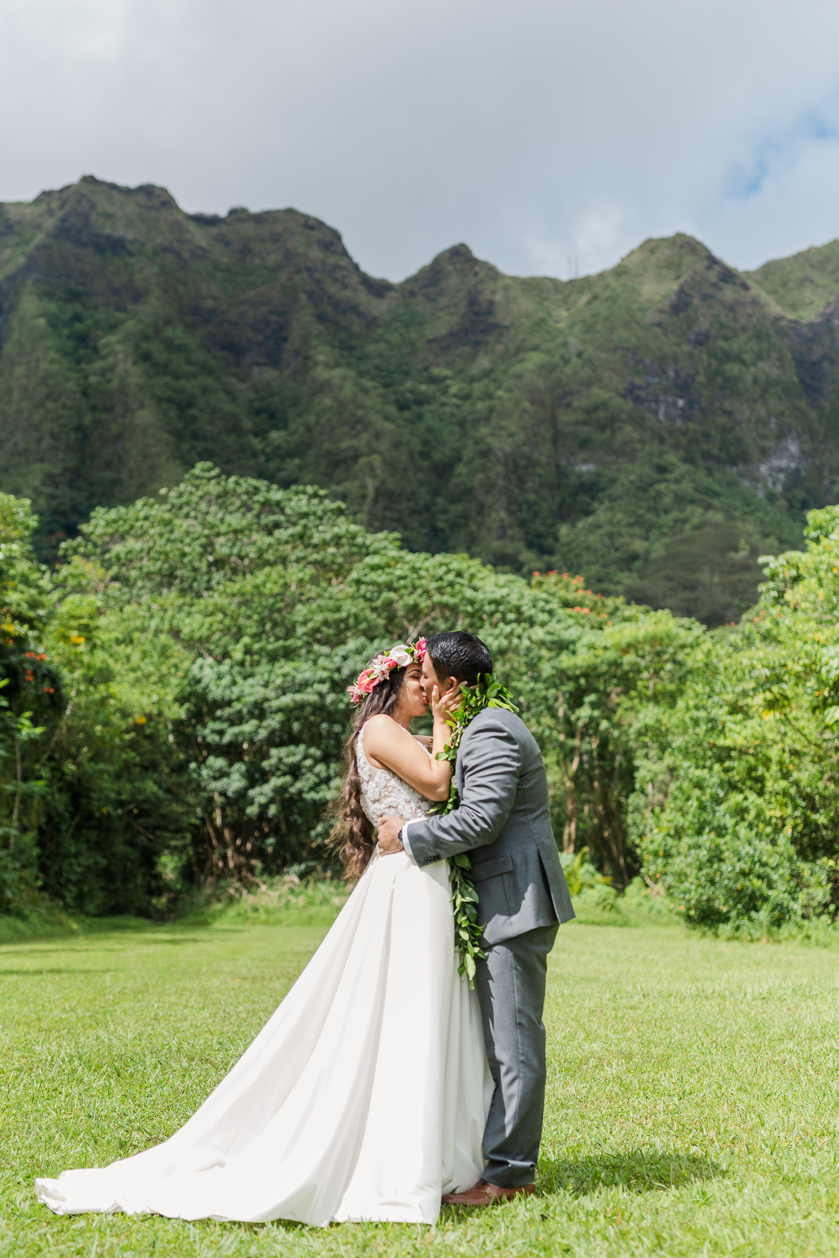 Newlywed couple sharing a second kiss wedding moment outdoors in a lush Hawaiian garden with mountains in the background. The bride in a white gown and groom in a gray suit celebrate their wedding day surrounded by tropical greenery.