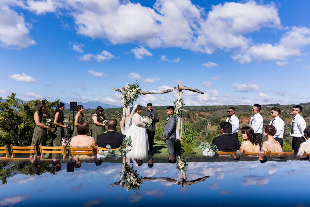 Bride and groom exchange vows beneath a driftwood floral arch at an outdoor Oahu wedding ceremony, reflected in an infinity pool with a dramatic blue sky and mountain ridgeline backdrop, photographed by Blessed Creatives
