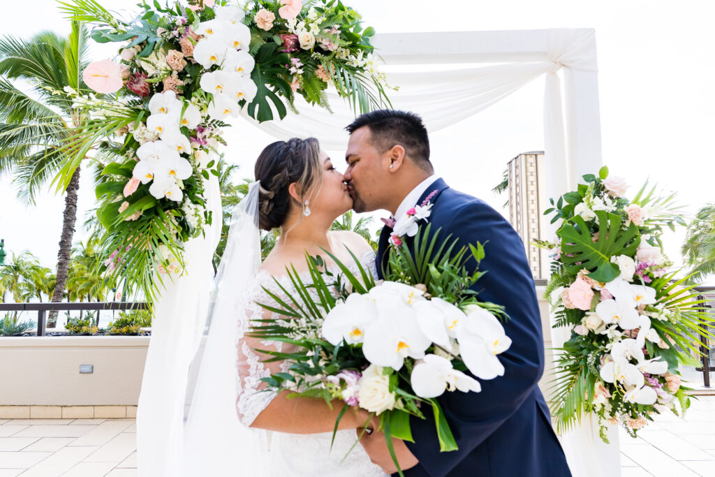 A bride and groom kiss as the bride holds a large white bouquet from a Hawaiian flower shop.