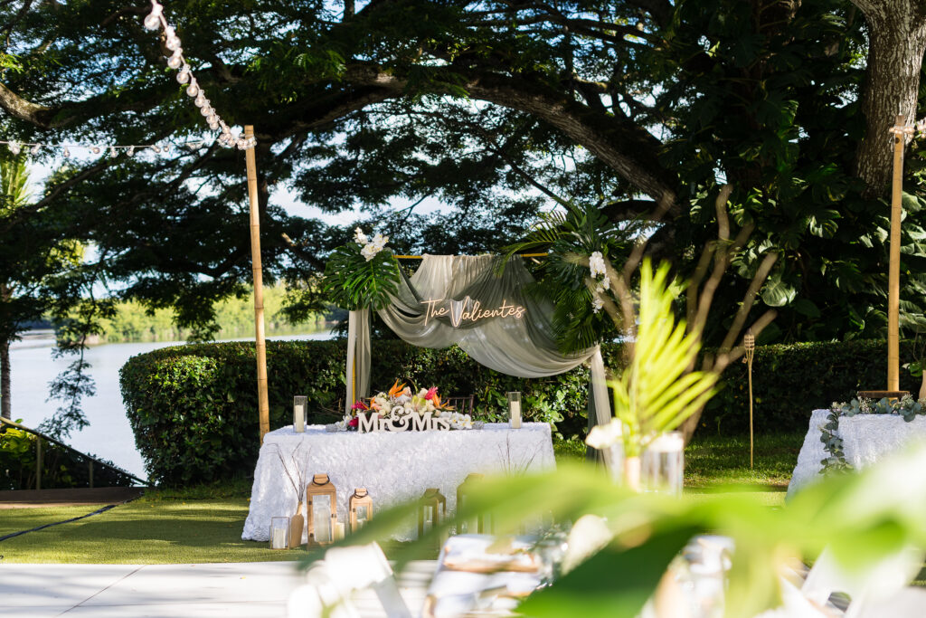Outdoor wedding reception set up in Hawaii, held under trees with tables and decorations, located next to a scenic body of water.