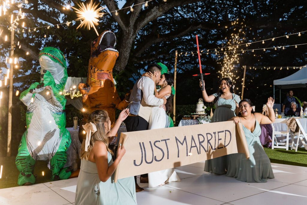 Bride and groom kiss at the center of the dance floor as their bridal party surrounds them, dancing with a “Just Married” sign and wearing playful dinosaur costumes.