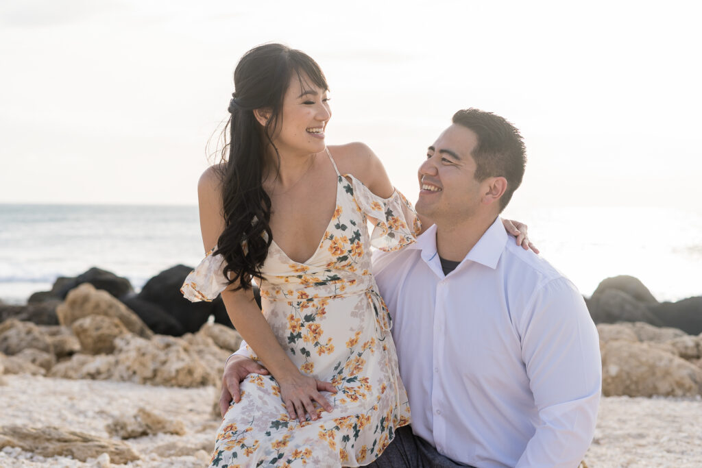 Engaged couple pose on a Hawaiian beach as the bride sits on the groom’s lap.