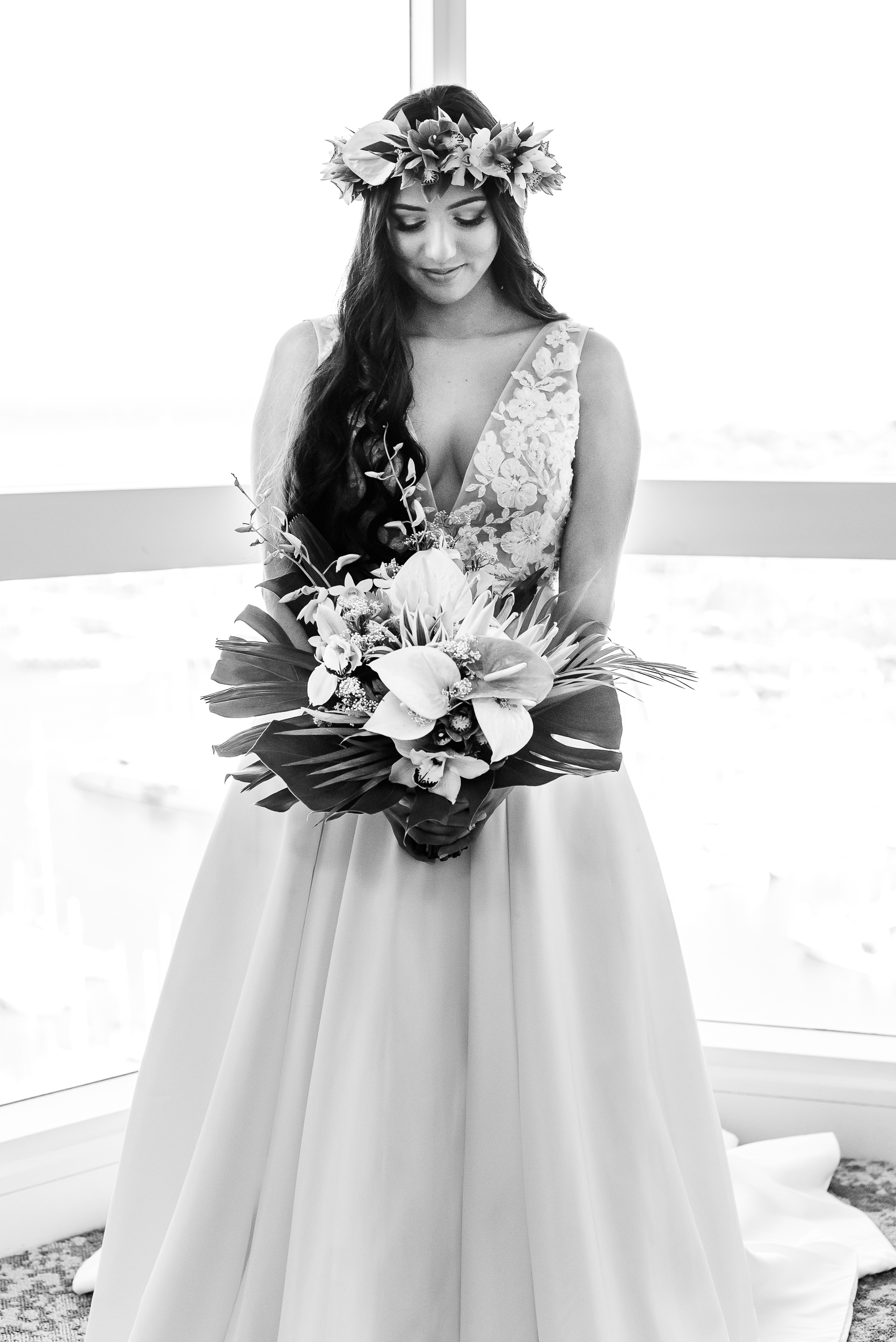 Bride standing next to tall windows at her wedding venue. She is looking down at her bouquet before her wedding.