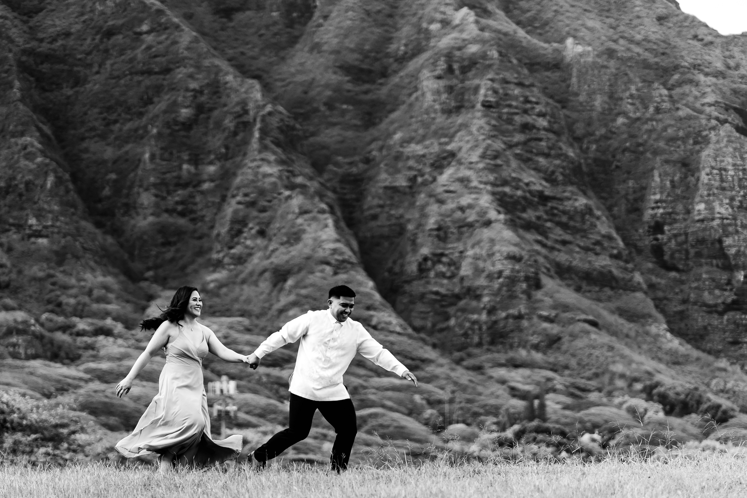 A newly married couple runs hand in hand in Hawaii with dramatic rock formations in the background.