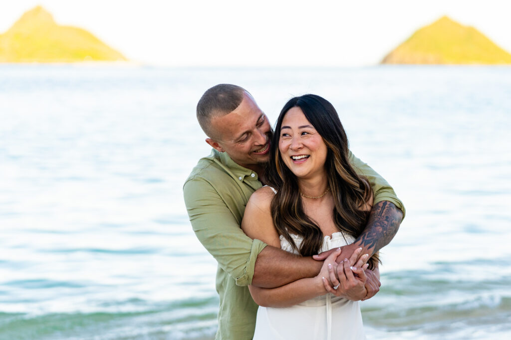 Joyful couple laughing together by the ocean with island views in the background during their Hawaii engagement photos.