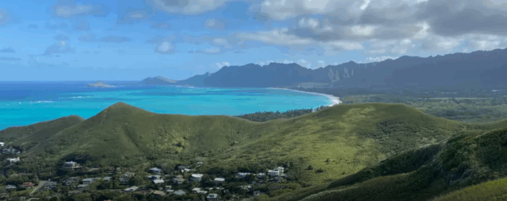 A scenic view from the Lanikai Pillbox Trail in Oahu, Hawaii, showing a dirt hiking path winding up a green hillside toward a small military pillbox. The bright blue ocean and Mokulua Islands are visible in the background under a clear sky.