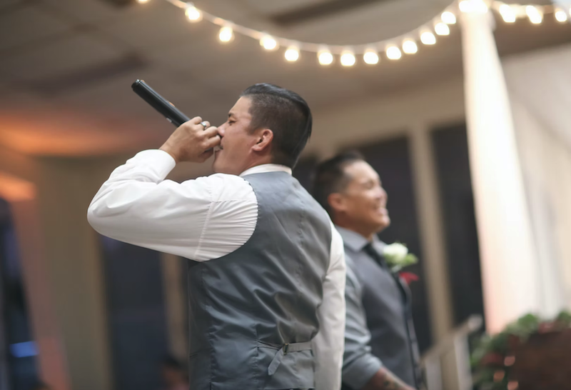 From Above Entertainment DJ and MC performing with a microphone during a wedding reception on Oahu under warm string lights.