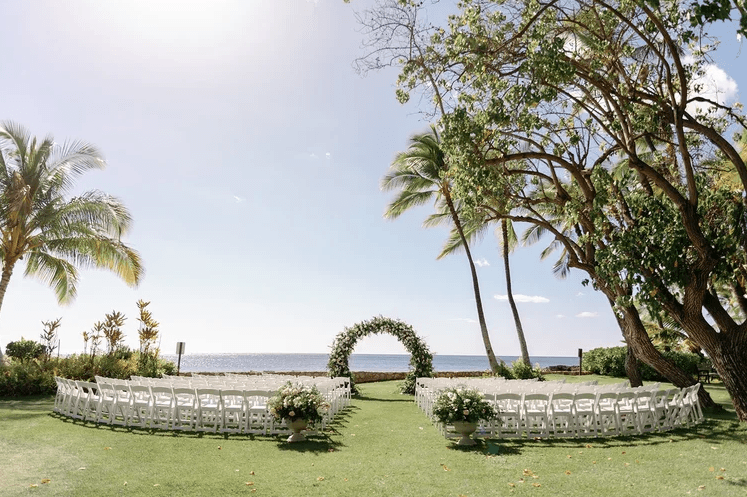 Oceanfront Hawaii wedding ceremony coordinated by Esselle Weddings featuring white chairs, floral arrangements, a greenery-covered arch, palm trees, and an ocean backdrop.