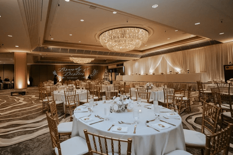 Elegant ballroom wedding coordinated by Esselle Weddings featuring white reception tables, gold chairs, white floral centerpieces, draped walls, and a large crystal chandelier.