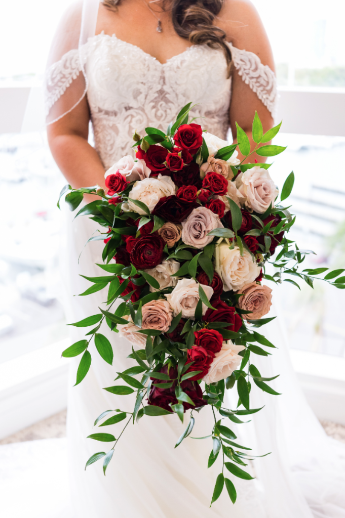 Bride holding a large cascading wedding bouquet of crimson, blush, and mauve roses with trailing greenery against a lace off-shoulder gown