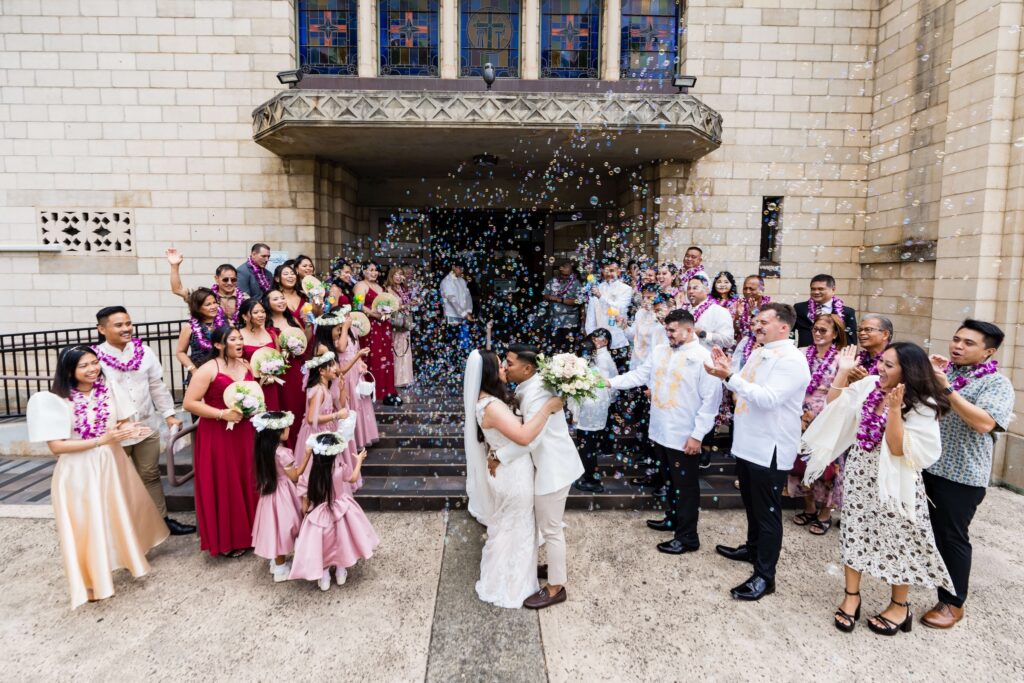 Newlywed couple sharing a second kiss wedding moment outside a church as family and friends celebrate with bubbles, smiles, and applause. The bride and groom stand surrounded by their wedding party and guests wearing traditional Hawaiian leis during their joyful ceremony exit.