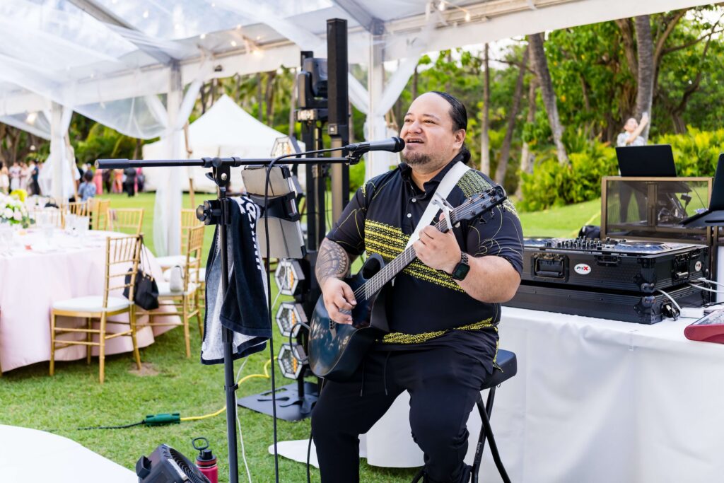 Live acoustic guitarist performing at an outdoor tented Hawaii wedding reception with gold chiavari chairs and tropical garden backdrop
